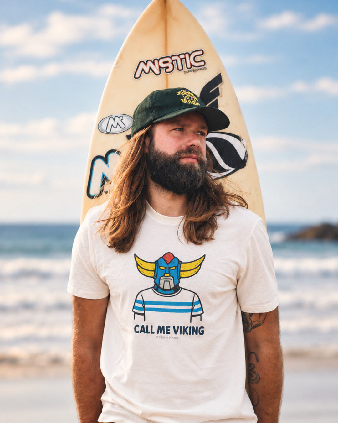 Homme portant un t-shirt blanc Ocean Park imprimé Call Me Viking, avec une casquette verte et une planche de surf, sur une plage bretonne.