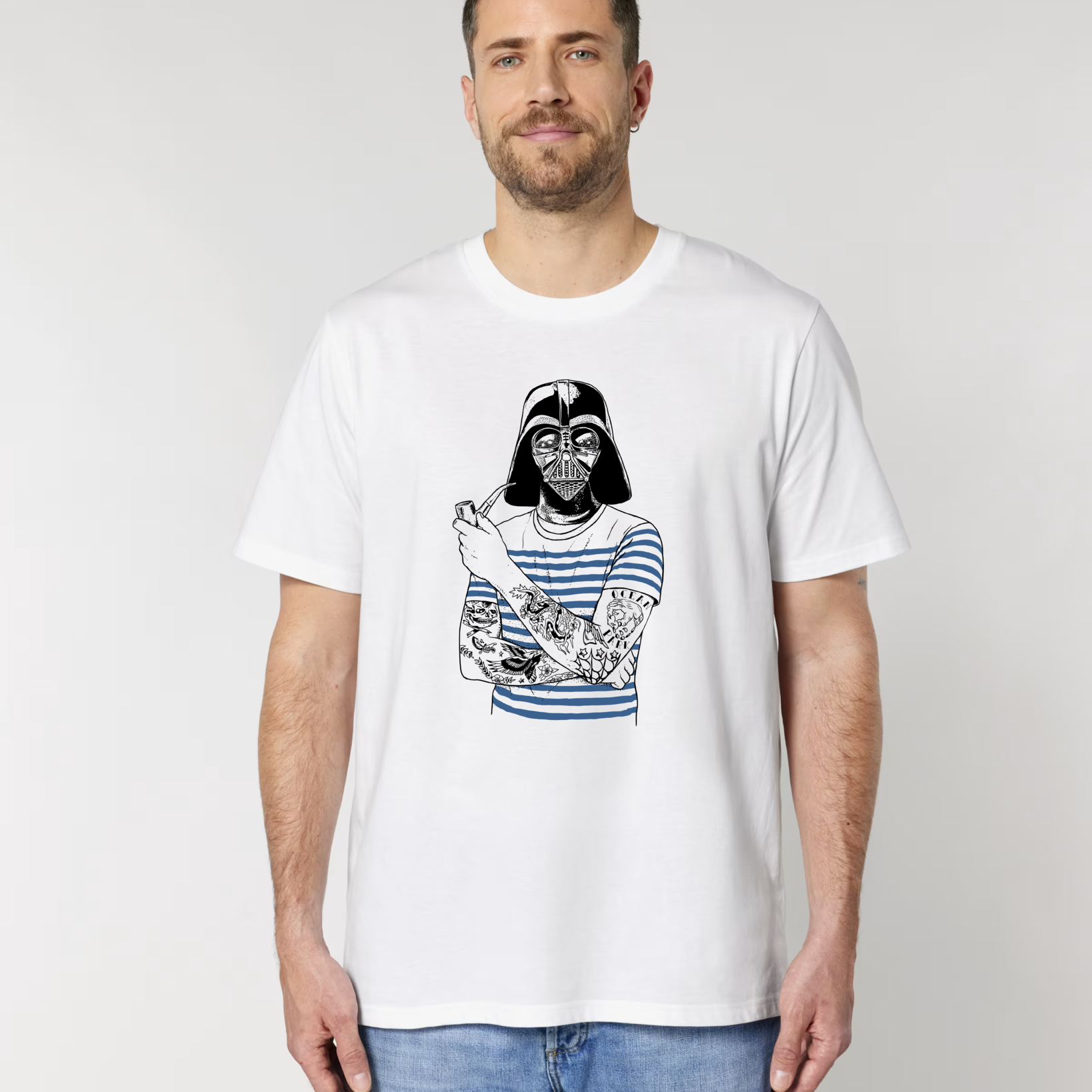 Un homme barbu de face portant un t-shirt blanc avec une illustration parodique de Dark Vador portant une marinière bleue, fumant la pipe et ayant les bras tatoués.