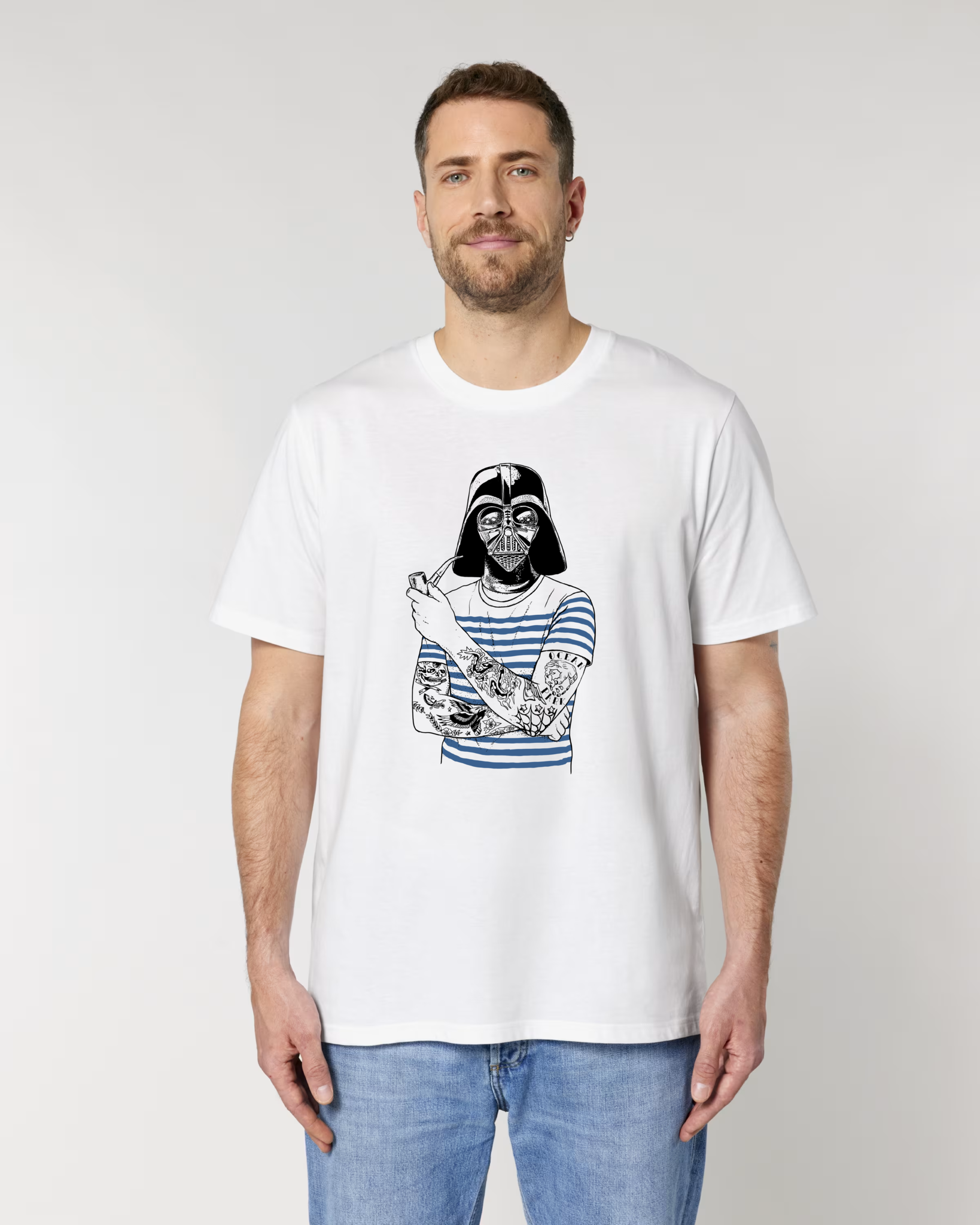 Un homme barbu de face portant un t-shirt blanc avec une illustration parodique de Dark Vador portant une marinière bleue, fumant la pipe et ayant les bras tatoués.
