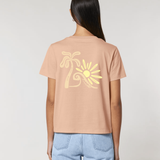 Vue de dos d'une femme portant un t-shirt couleur pêche avec une grande illustration jaune pâle représentant un palmier, une vague et un soleil stylisés.