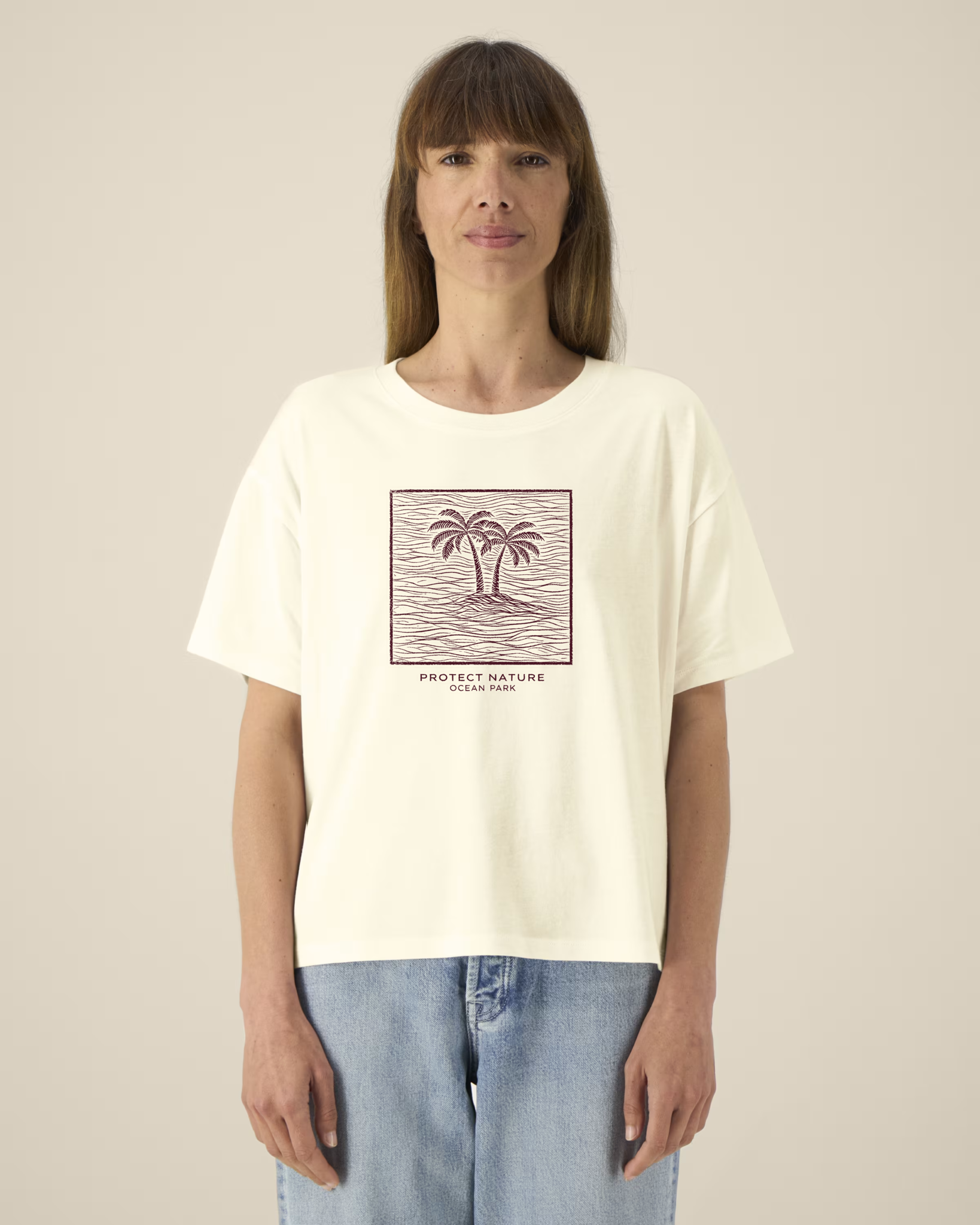 Femme de face portant un t-shirt de couleur crème avec une illustration centrale bordeaux représentant deux palmiers sur une île entourée de vagues dans un cadre carré, avec l'inscription "PROTECT NATURE OCEAN PARK".