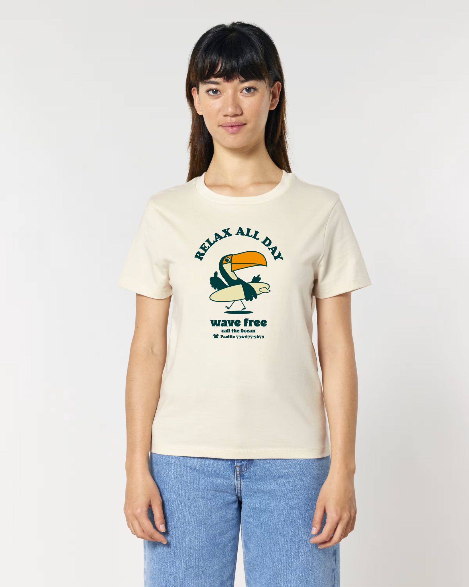 Femme de face portant un t-shirt de couleur blanc cassé naturel avec une illustration centrale d'un toucan tenant une planche de surf, entouré des textes "RELAX ALL DAY" et "wave free call the Ocean".