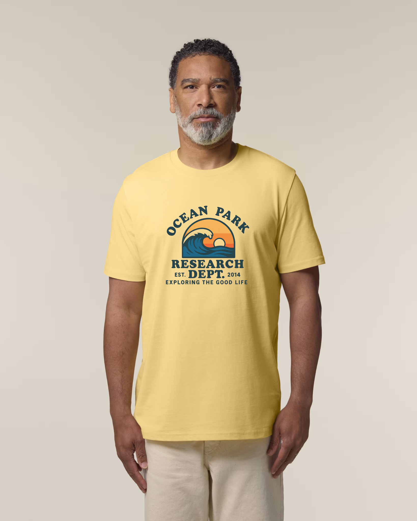 Un homme d'âge mûr avec une barbe grise porte un t-shirt jaune "viva yellow".
Le visuel central montre une vague bleue devant un soleil couchant orange, entourée des textes "OCEAN PARK RESEARCH DEPT. EST. 2014 EXPLORING THE GOOD LIFE".
