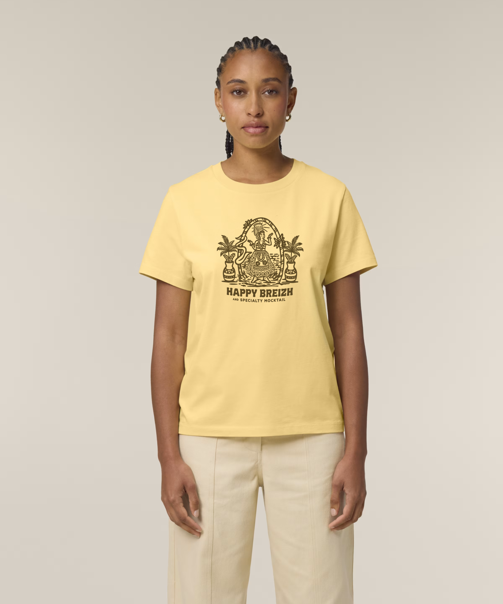 Femme de face portant un t-shirt jaune clair avec une illustration marron représentant une bigoudène dans un décor de plage tropicale avec palmiers et l'inscription "HAPPY BREIZH".