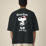Vue de dos d'un homme portant un t-shirt noir oversize avec une grande illustration d'un chien de type cartoon (Snoopy) déguisé en rappeur avec bandana rouge, lunettes de soleil, tresse et baskets, surmonté du texte blanc "Ocean Park".