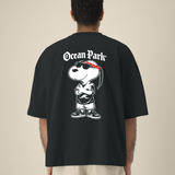 Vue de dos d'un homme portant un t-shirt noir oversize avec une grande illustration d'un chien de type cartoon (Snoopy) déguisé en rappeur avec bandana rouge, lunettes de soleil, tresse et baskets, surmonté du texte blanc "Ocean Park".