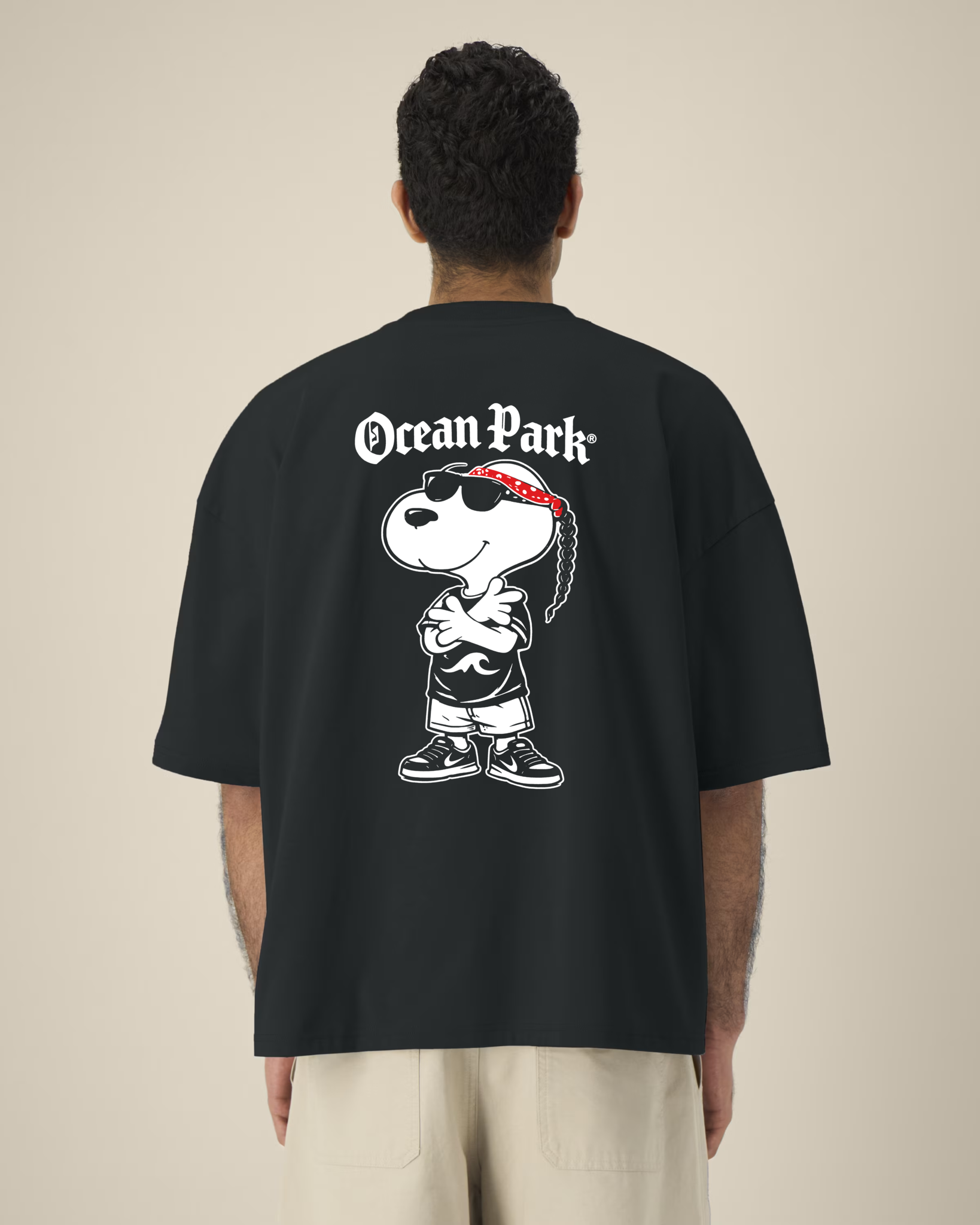 Vue de dos d'un homme portant un t-shirt noir oversize avec une grande illustration d'un chien de type cartoon (Snoopy) déguisé en rappeur avec bandana rouge, lunettes de soleil, tresse et baskets, surmonté du texte blanc "Ocean Park".