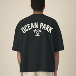 Vue de dos d'un homme portant un t-shirt noir oversize avec une grande inscription blanche "OCEAN PARK" en arc de cercle, présentant un effet de texture ruban adhésif, surmontant une illustration de deux palmiers.