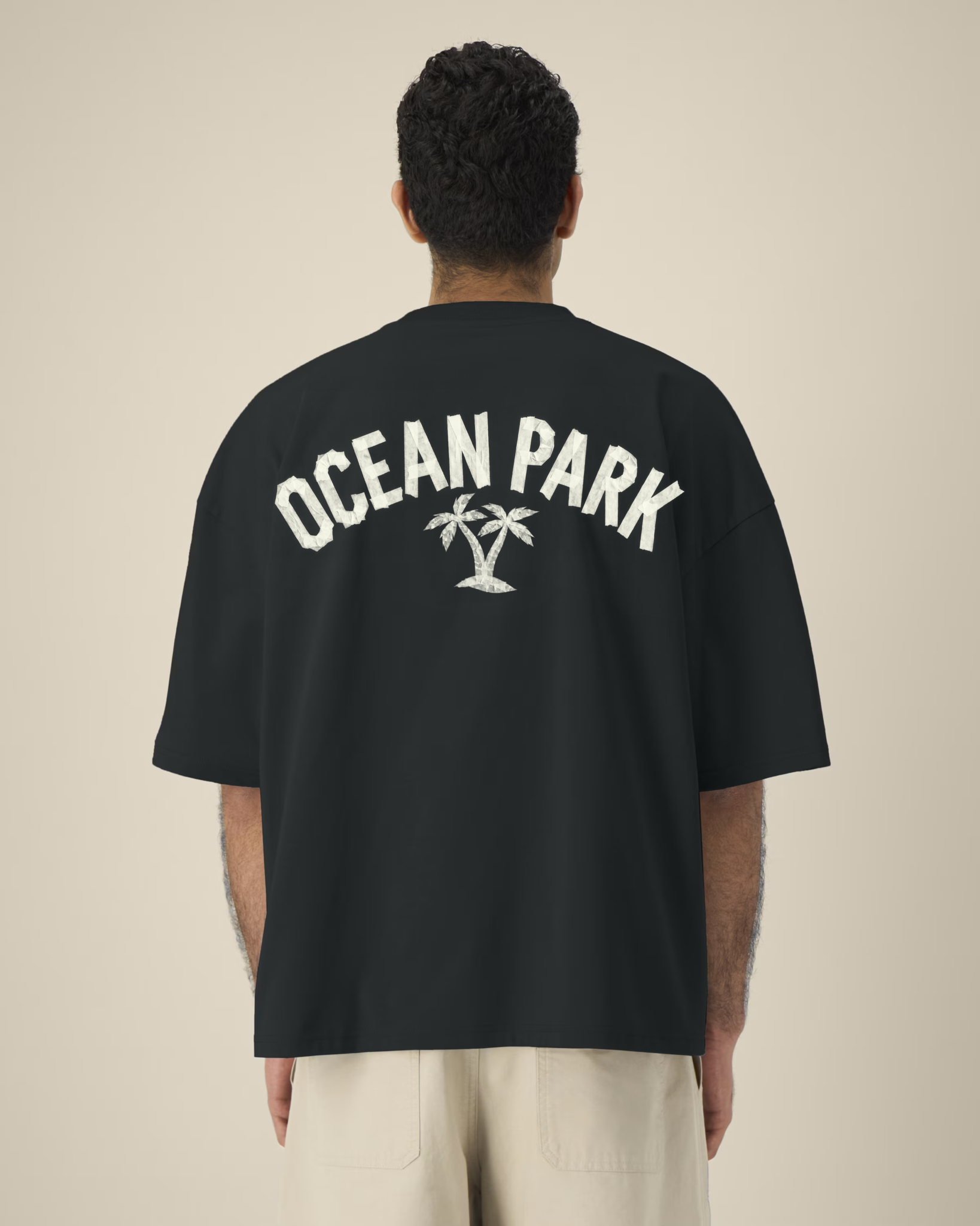 Vue de dos d'un homme portant un t-shirt noir oversize avec une grande inscription blanche "OCEAN PARK" en arc de cercle, présentant un effet de texture ruban adhésif, surmontant une illustration de deux palmiers.