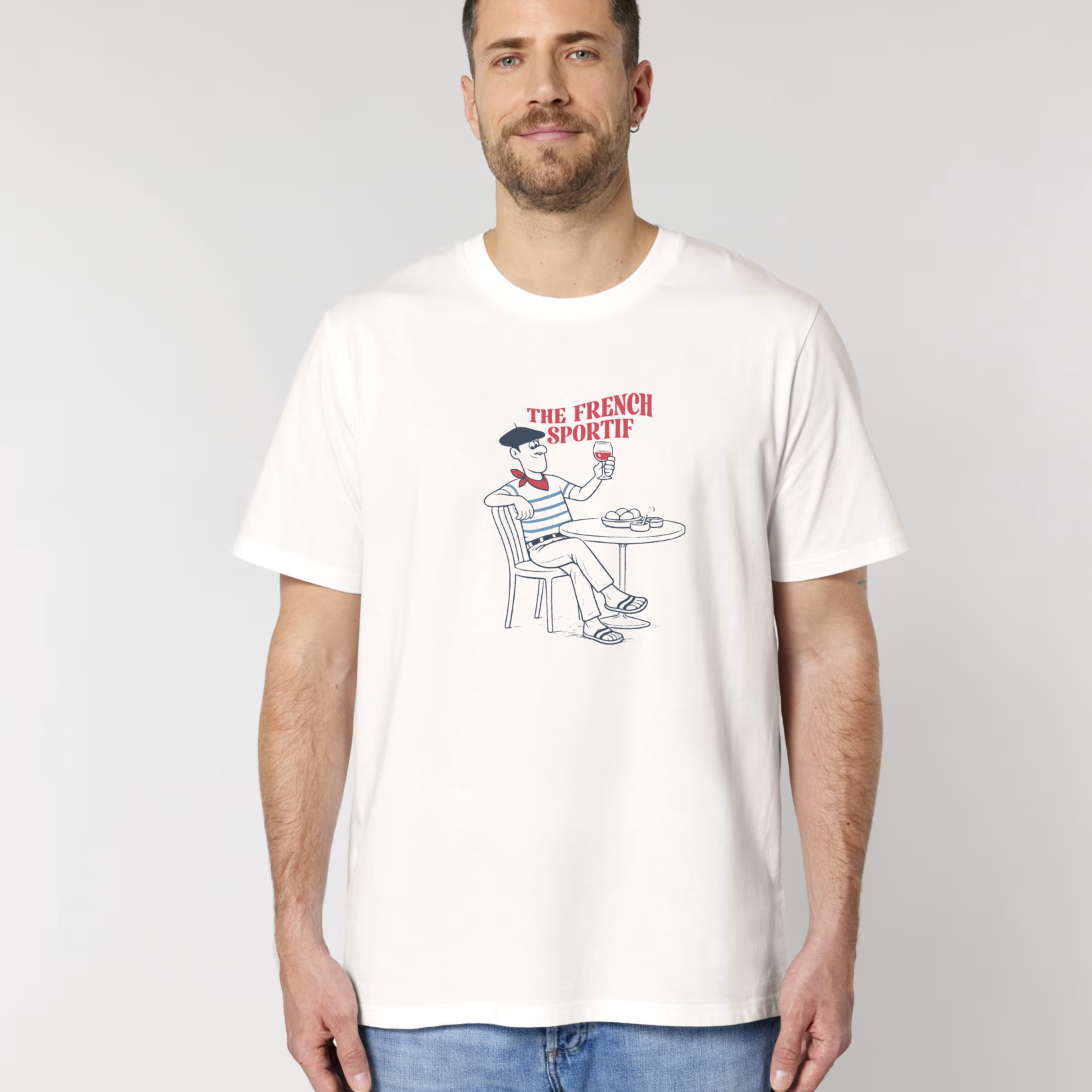 Homme portant un t-shirt blanc cassé avec une illustration centrale d'un homme à une terrasse de café, portant un béret et une marinière, tenant un verre de vin rouge sous le texte "THE FRENCH SPORTIF".