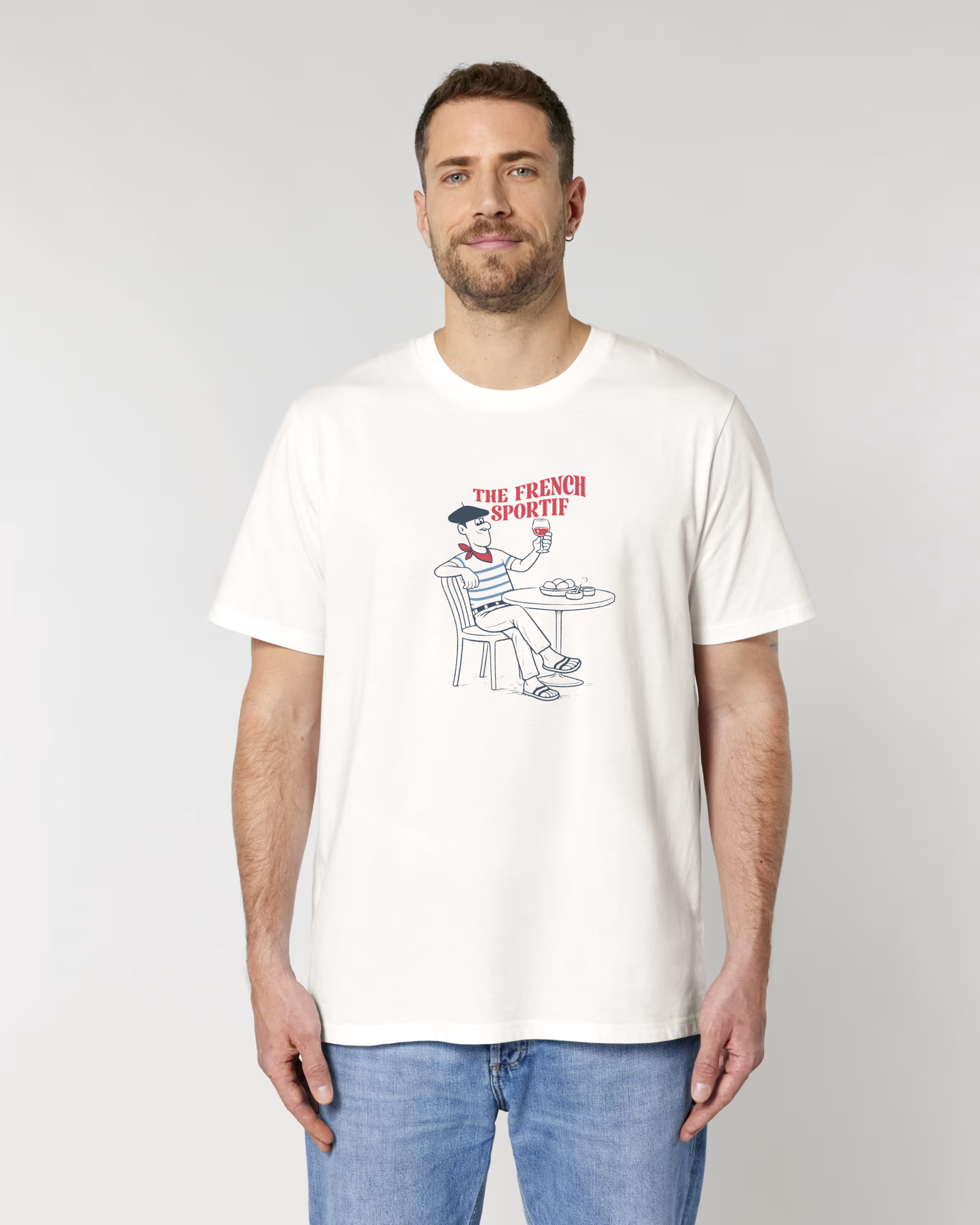 Homme portant un t-shirt blanc cassé avec une illustration centrale d'un homme à une terrasse de café, portant un béret et une marinière, tenant un verre de vin rouge sous le texte "THE FRENCH SPORTIF".