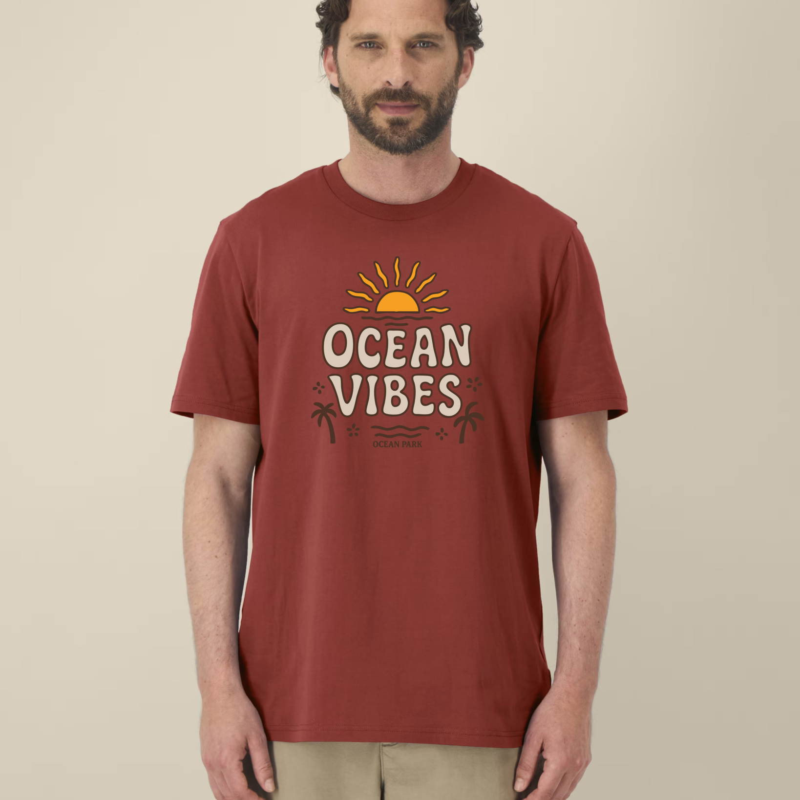 Homme de face portant un t-shirt de couleur rouge terreux (earthy red) avec un graphisme central composé d'un soleil couchant jaune, du texte "OCEAN VIBES" en lettres beiges arrondies, et des silhouettes de palmiers marron.