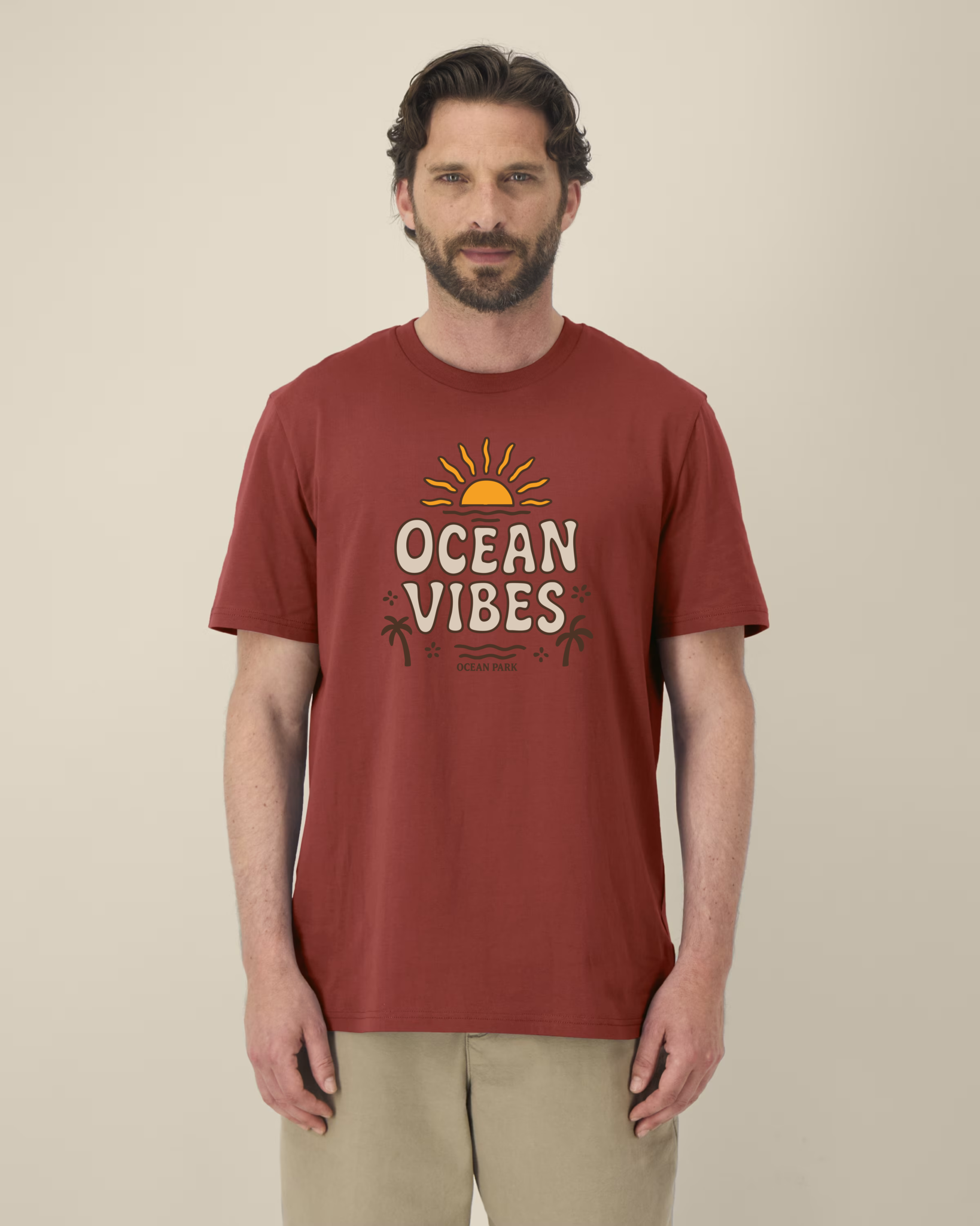 Homme de face portant un t-shirt de couleur rouge terreux (earthy red) avec un graphisme central composé d'un soleil couchant jaune, du texte "OCEAN VIBES" en lettres beiges arrondies, et des silhouettes de palmiers marron.