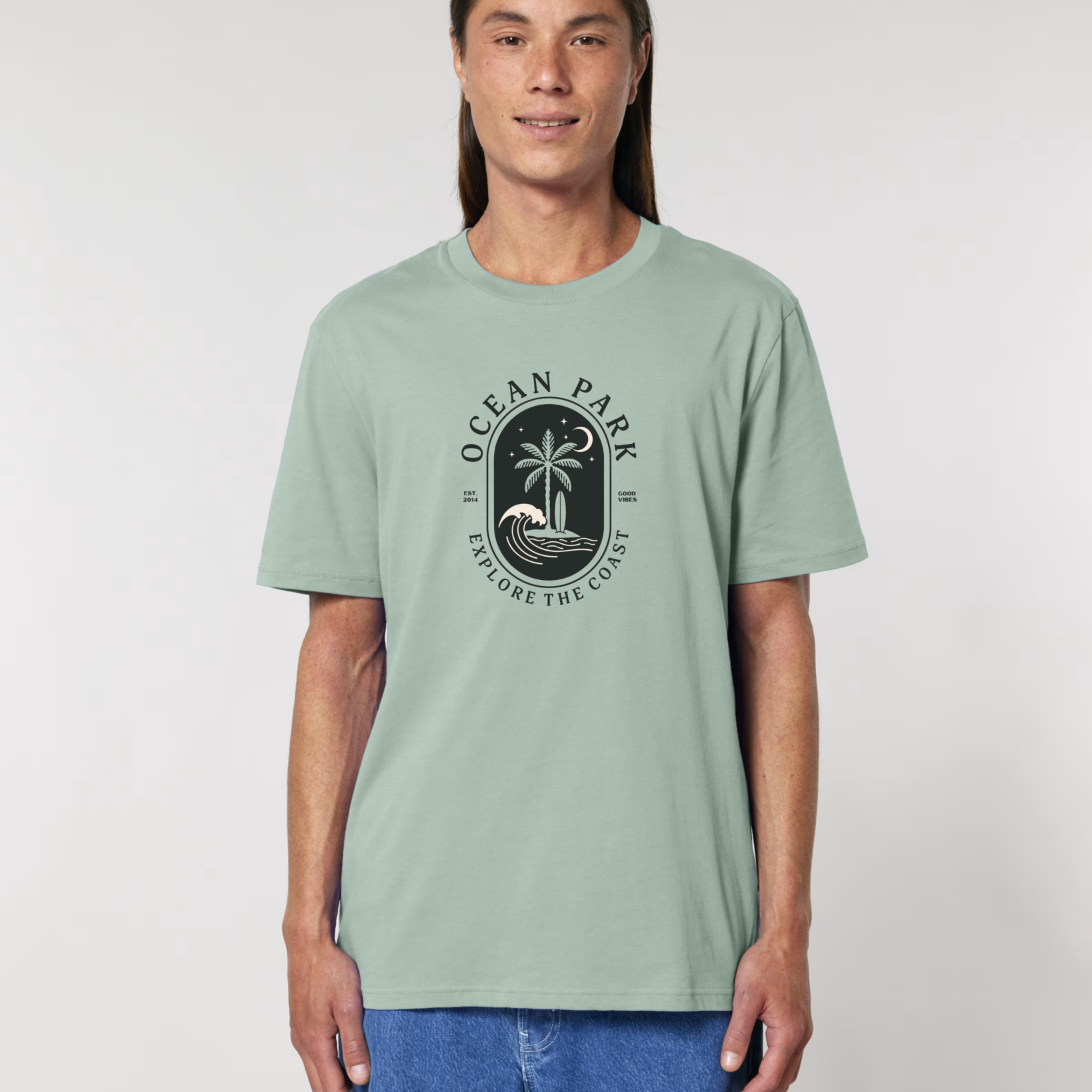 Homme de face portant un t-shirt de couleur vert aloe avec un graphisme central en forme de badge noir contenant un palmier, une planche de surf, une vague et un croissant de lune, entouré du texte "Ocean Park - Explore The Coast".