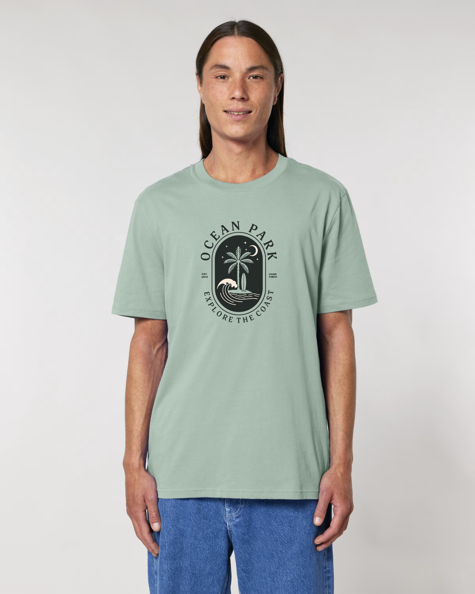Homme de face portant un t-shirt de couleur vert aloe avec un graphisme central en forme de badge noir contenant un palmier, une planche de surf, une vague et un croissant de lune, entouré du texte "Ocean Park - Explore The Coast".