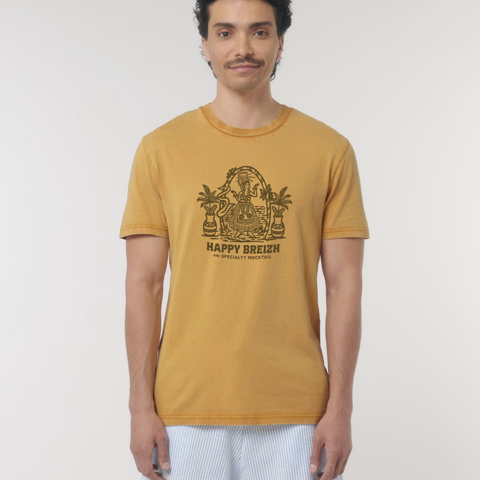 Homme portant un t-shirt vintage de couleur ocre doré avec une illustration marron représentant une femme en costume traditionnel breton (bigoudène) dans un cadre tropical avec palmiers, surmontant le texte "HAPPY BREIZH".