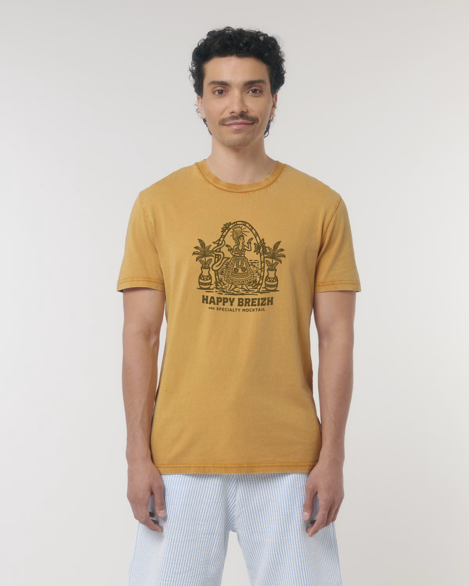 Homme portant un t-shirt vintage de couleur ocre doré avec une illustration marron représentant une femme en costume traditionnel breton (bigoudène) dans un cadre tropical avec palmiers, surmontant le texte "HAPPY BREIZH".