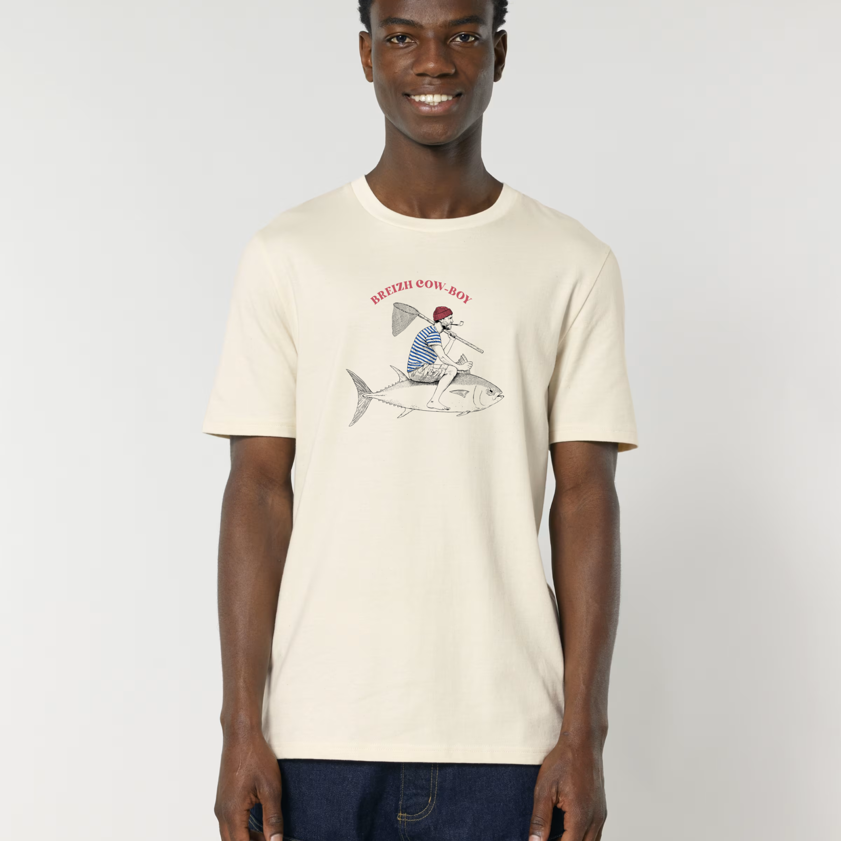 Un homme souriant portant un t-shirt de couleur crème (écru) avec une illustration d'un marin en marinière chevauchant un gros thon comme un cowboy, avec l'inscription rouge "BREIZH COW-BOY".