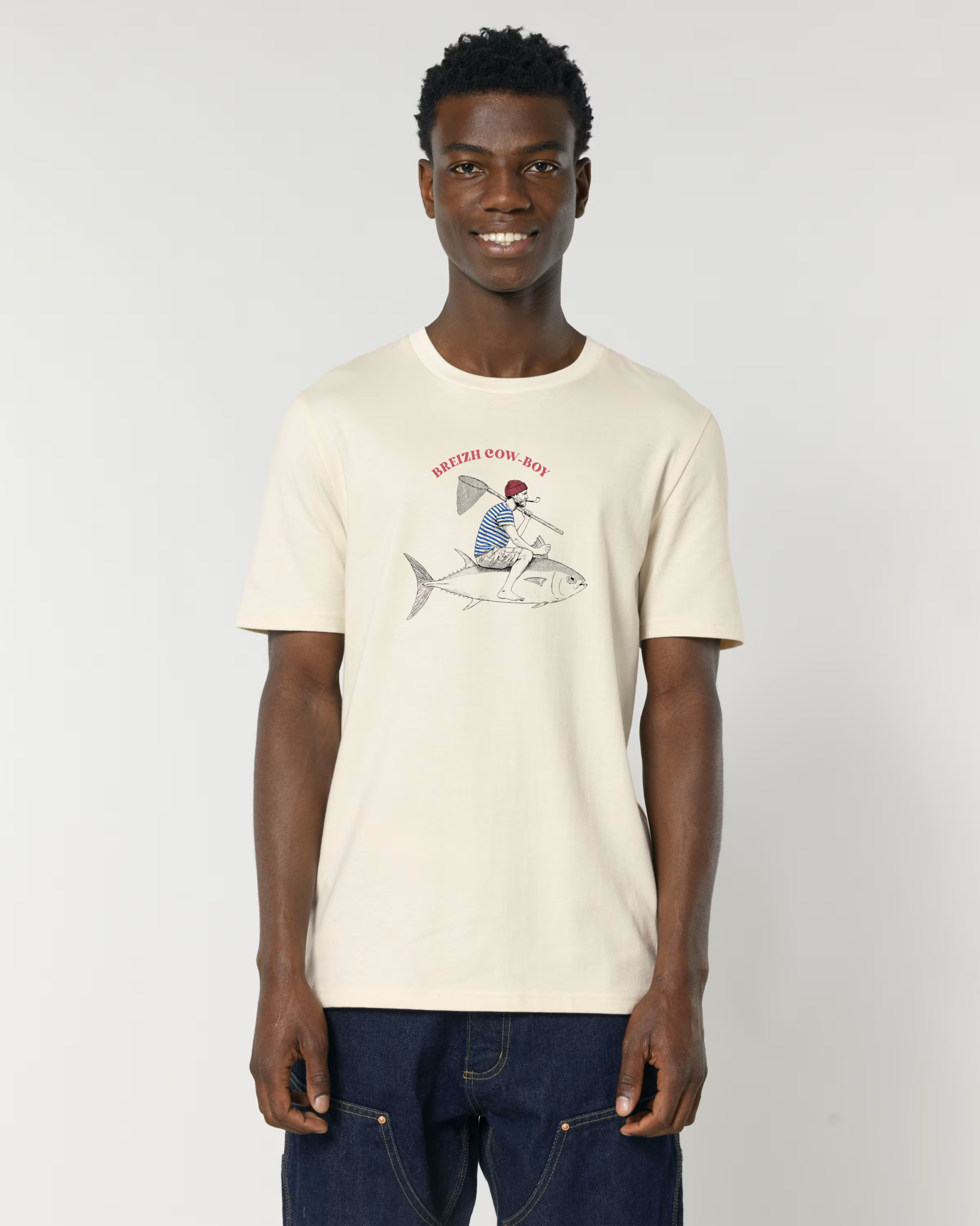 Un homme souriant portant un t-shirt de couleur crème (écru) avec une illustration d'un marin en marinière chevauchant un gros thon comme un cowboy, avec l'inscription rouge "BREIZH COW-BOY".